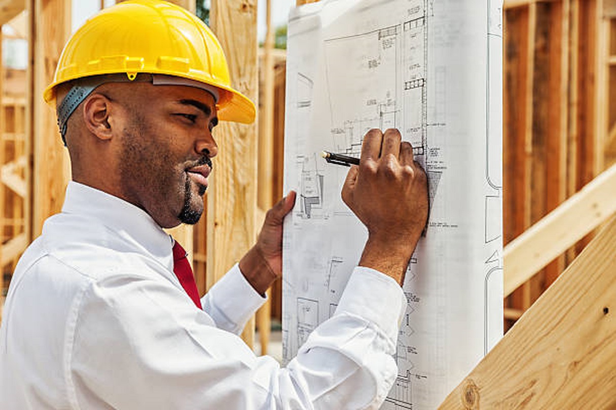Photo of an architect at construction site, holding building plans under his arm, making notes with mechanical pencil.